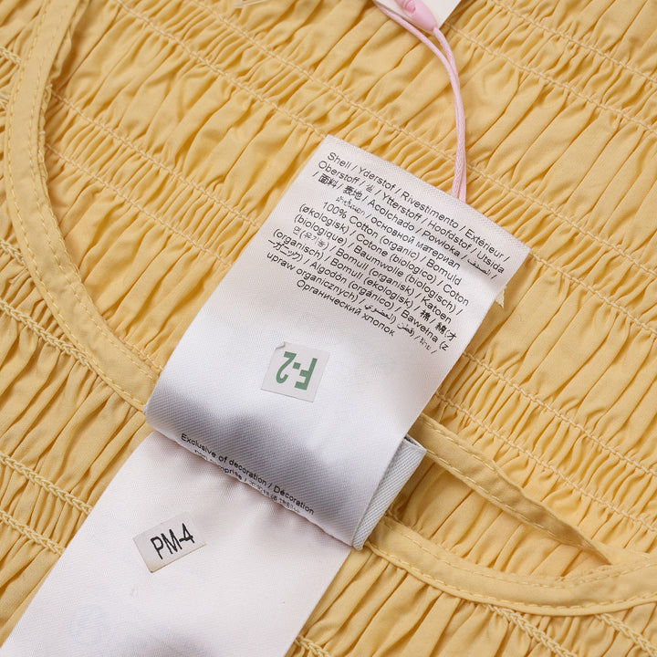 Ganni butter yellow sleeveless smocked peplum blouse with all-over shirring, side tie bows, and flounced hem, photographed flat on white background