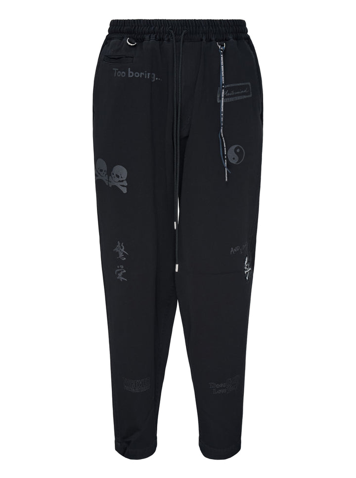Mastermind Doodle Print Easy Pants in black with all-over graphic prints including skull-and-crossbones, yin-yang symbol, kanji script, and hand-scrawled text, featuring an elasticated drawstring waistband and tapered leg