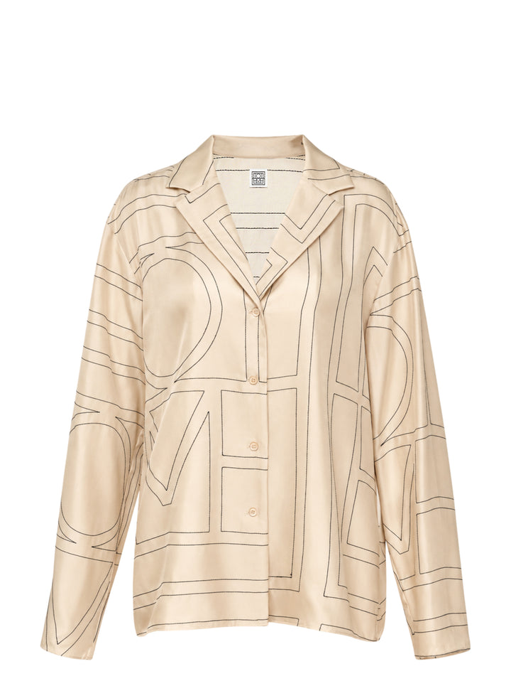Totême Monogram Silk Pyjama Top in cream with dark outlined oversized logo lettering, notched lapel collar, button-through front, and long sleeves