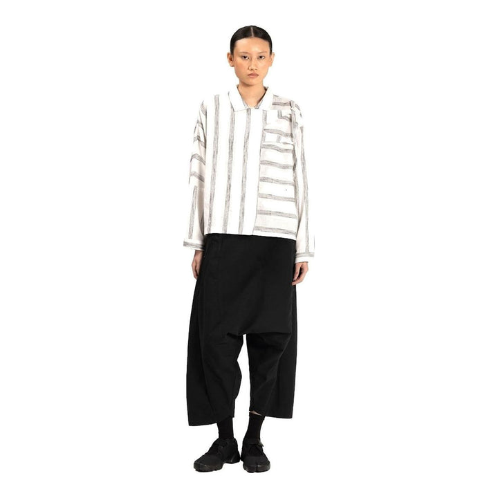 Model wearing Anaabu Asymmetrical Batwing Top in black textured fabric with an open collar, asymmetric front placket, patch chest pocket, and long batwing sleeves, styled with dark drop-crotch trousers and brown mules - packshot