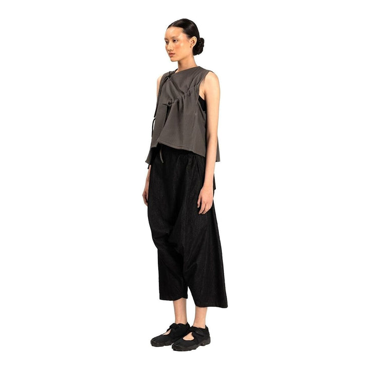 Model wearing the Anaabu Asymmetrical Vest in black, featuring an asymmetric neckline, diagonal panelling, and a draped fringe tie at the shoulder, styled with wide-leg black trousers and dark leather sandals. - packshot