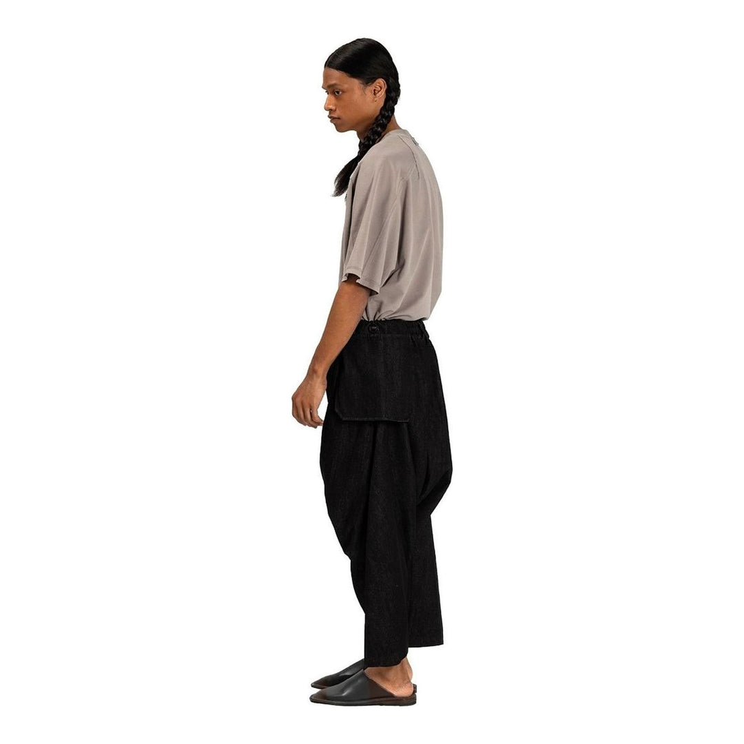 Model wearing Anaabu Dropped Culotte Pants in black — wide-leg drop-crotch trousers with an asymmetric draped skirt overlay panel, styled with a black crop top, frayed bucket hat, and tabi sneakers - packshot
