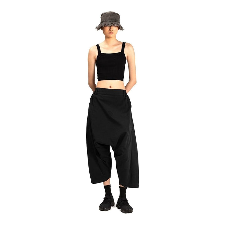 Model wearing Anaabu Dropped Culotte Pants in black — wide-leg drop-crotch trousers with an asymmetric draped skirt overlay panel, styled with a black crop top, frayed bucket hat, and tabi sneakers - packshot