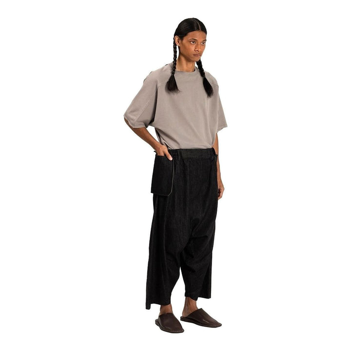 Model wearing Anaabu Dropped Culotte Pants in black — wide-leg drop-crotch trousers with an asymmetric draped skirt overlay panel, styled with a black crop top, frayed bucket hat, and tabi sneakers - packshot