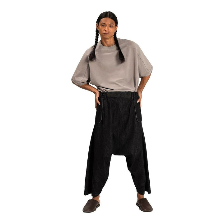 Model wearing Anaabu Dropped Culotte Pants in black — wide-leg drop-crotch trousers with an asymmetric draped skirt overlay panel, styled with a black crop top, frayed bucket hat, and tabi sneakers - packshot