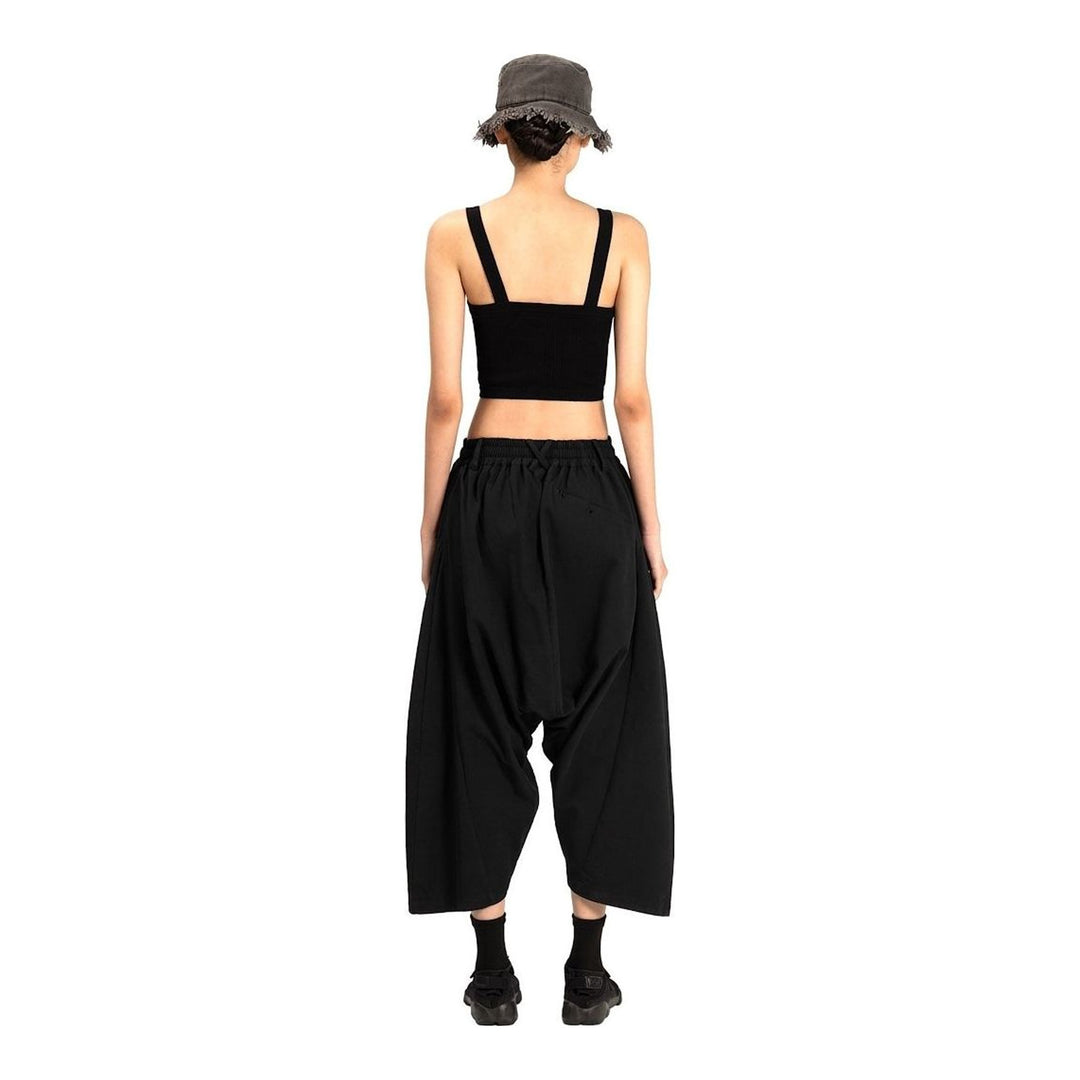 Model wearing Anaabu Dropped Culotte Pants in black — wide-leg drop-crotch trousers with an asymmetric draped skirt overlay panel, styled with a black crop top, frayed bucket hat, and tabi sneakers - packshot