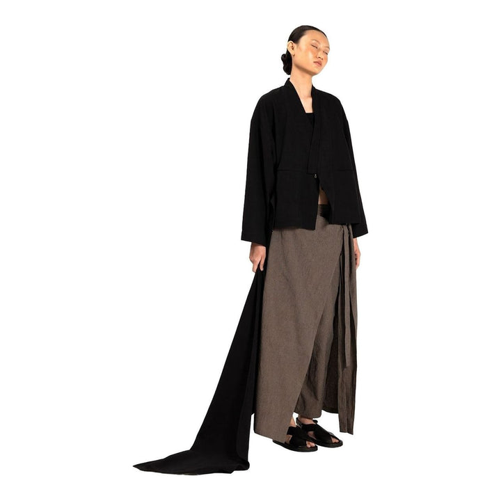 Model wearing the Anaabu Shawl Jacket in black — a draped wrap-style jacket with oversized shawl collar and self-tie belt, styled over a black turtleneck and olive wide-leg trousers - packshot