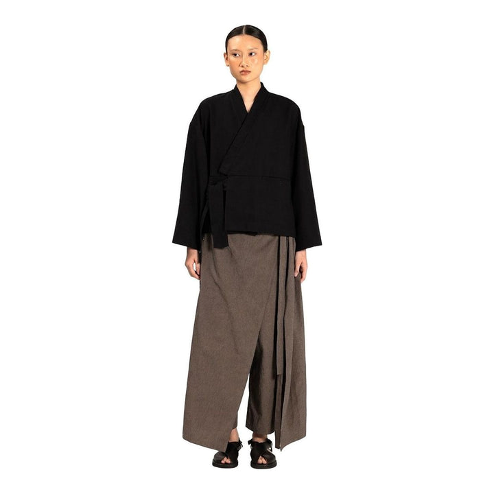 Model wearing the Anaabu Shawl Jacket in black — a draped wrap-style jacket with oversized shawl collar and self-tie belt, styled over a black turtleneck and olive wide-leg trousers - packshot