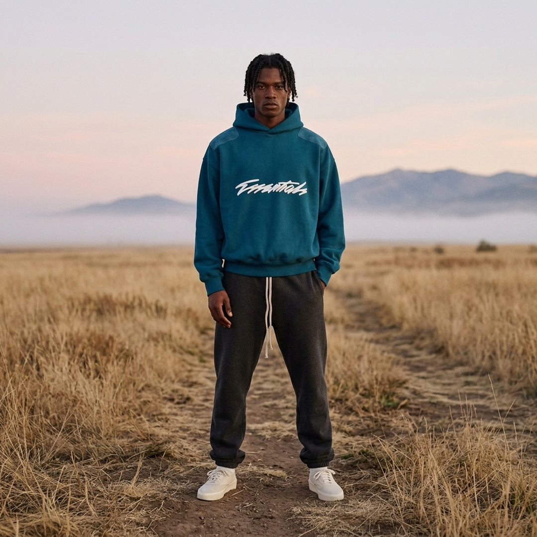 Fear of God Essentials Classic Raglan Hoodie in deep teal with white script logo across the chest and tonal textured shoulder panels - lifestyle